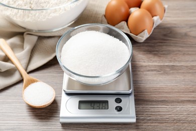Photo of Digital kitchen scale with bowl of sugar and spoon on wooden table, closeup