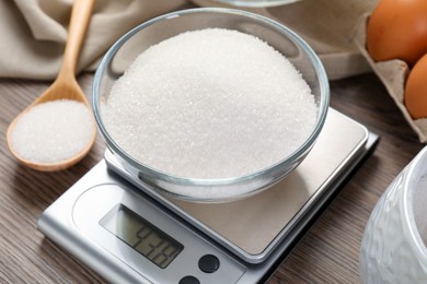 Photo of Digital kitchen scale with bowl of sugar on wooden table, closeup