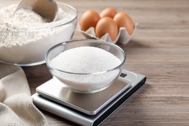 Photo of Digital kitchen scale with bowl of sugar and other products on wooden table, closeup