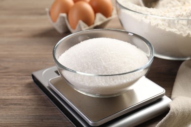 Photo of Digital kitchen scale with bowl of sugar and other products on wooden table, closeup