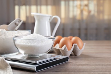 Photo of Digital kitchen scale with bowl of sugar and other products on wooden table indoors, closeup. Space for text