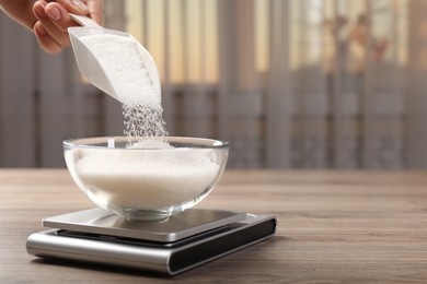 Photo of Woman adding sugar into bowl and digital kitchen scale at wooden table indoors, closeup. Space for text