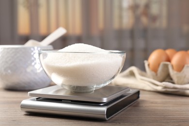 Photo of Digital kitchen scale with bowl of sugar on wooden table indoors, closeup