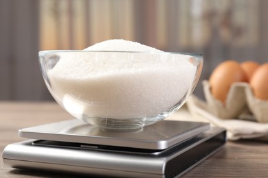 Photo of Digital kitchen scale with bowl of sugar on wooden table indoors, closeup