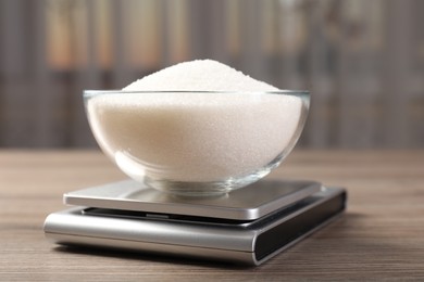Photo of Digital kitchen scale with bowl of sugar on wooden table indoors, closeup