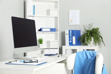 Photo of Doctor's workplace. Computer monitor, clipboard, stethoscope and glasses on white table in medical office