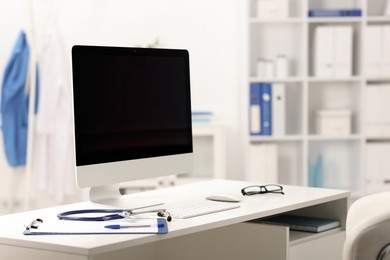 Photo of Doctor's workplace. Computer monitor, keyboard, mouse, glasses, stethoscope and stationery on white table in medical office