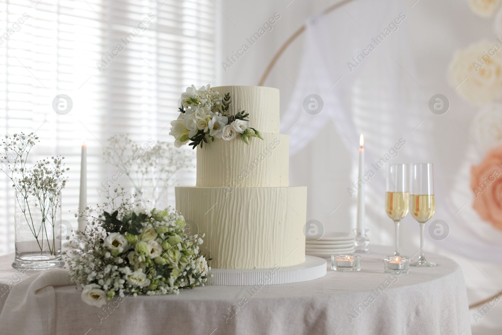 Photo of Delicious wedding cake with floral decor, bouquet and glasses of champagne on table indoors