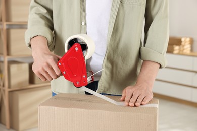 Photo of Man using dispenser with adhesive tape to seal cardboard box indoors, closeup