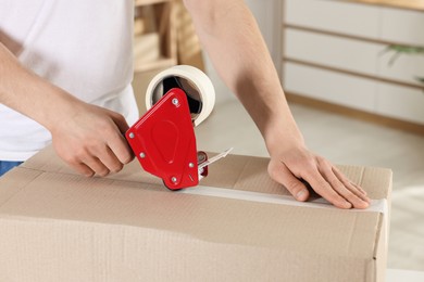 Photo of Man using dispenser with adhesive tape to seal cardboard box indoors, closeup