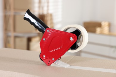 Photo of Dispenser with adhesive tape on cardboard box indoors, closeup