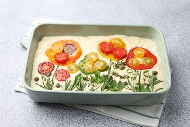 Photo of Raw focaccia with vegetables and spices in baking dish on light grey table, closeup