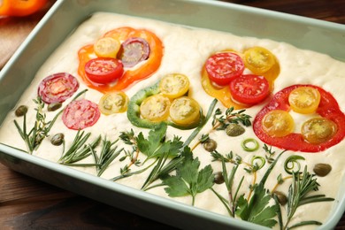 Photo of Raw focaccia with vegetables and spices in baking dish on wooden table, closeup