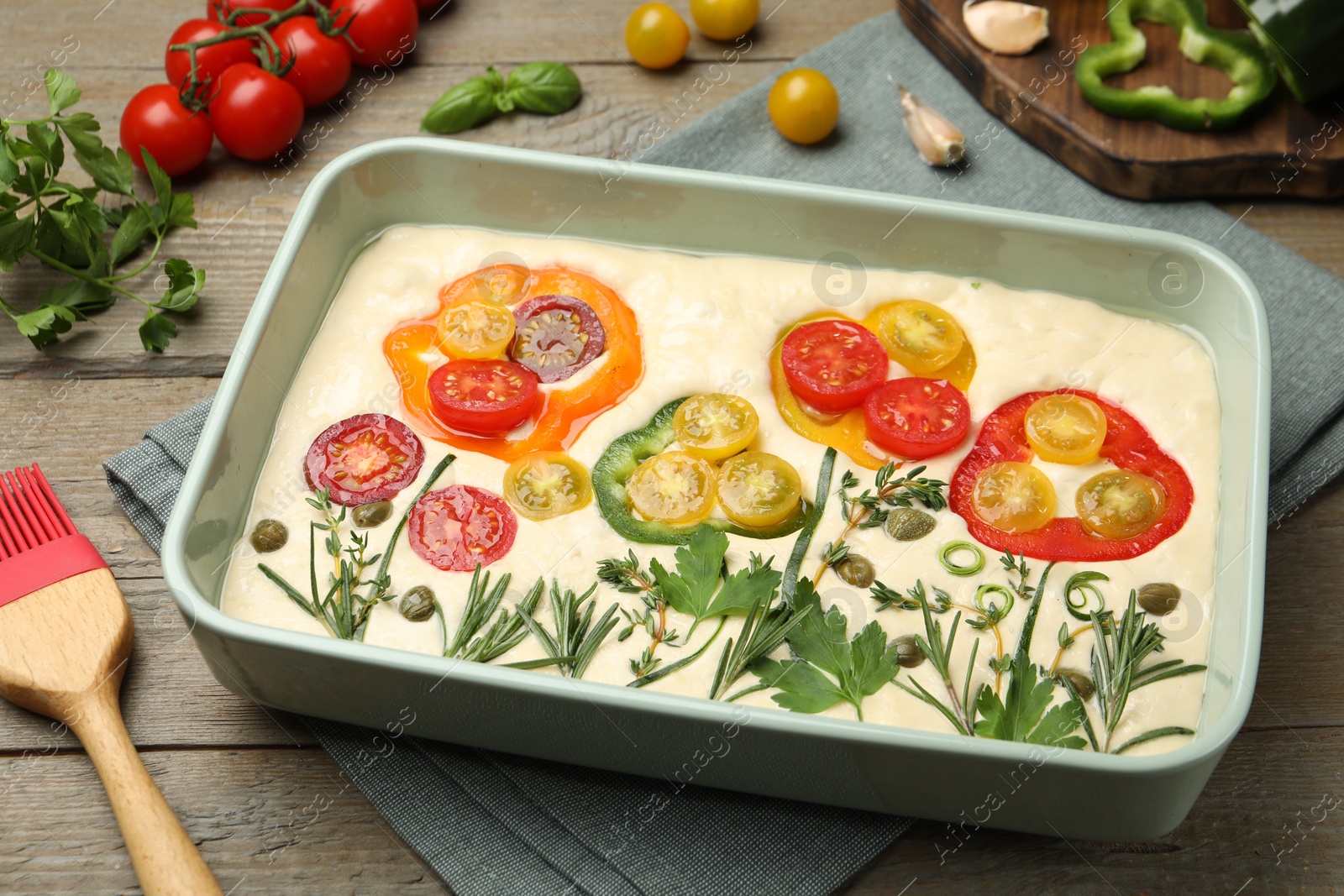 Raw focaccia with vegetables, spices and brush on wooden table, closeup Photo of Raw focaccia with vegetables, spices and brush on wooden table, closeup