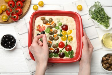 Photo of Woman adding basil to raw focaccia at white wooden table, top view