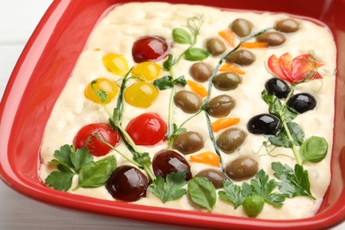 Photo of Raw focaccia with vegetables and olives in baking dish on table, closeup