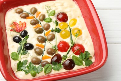 Photo of Raw focaccia with vegetables and olives in baking dish on white wooden table, above view