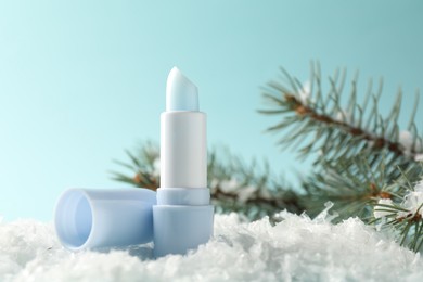 Photo of Moisturizing lip balm and fir tree branches on artificial snow against light blue background, closeup. Winter care