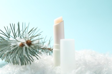 Photo of Moisturizing lip balm and fir tree branches on artificial snow against light blue background, closeup. Winter care