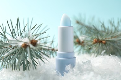Photo of Moisturizing lip balm and fir tree branches on artificial snow against light blue background, closeup. Winter care