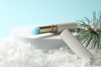 Photo of Moisturizing lip balm and fir tree branches on artificial snow against light blue background, closeup. Winter care