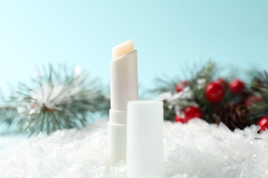 Photo of Moisturizing lip balm and Christmas decor on artificial snow against light blue background, closeup. Winter care