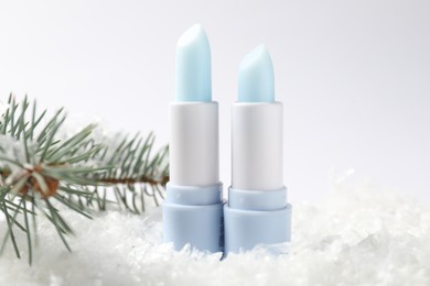 Photo of Moisturizing lip balms and fir tree branches on artificial snow against white background, closeup. Winter care
