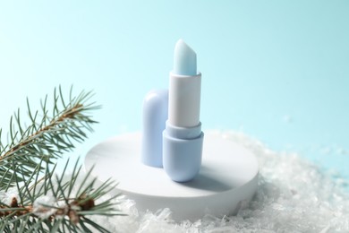 Photo of Moisturizing lip balm and fir tree branches on artificial snow against light blue background, closeup. Winter care