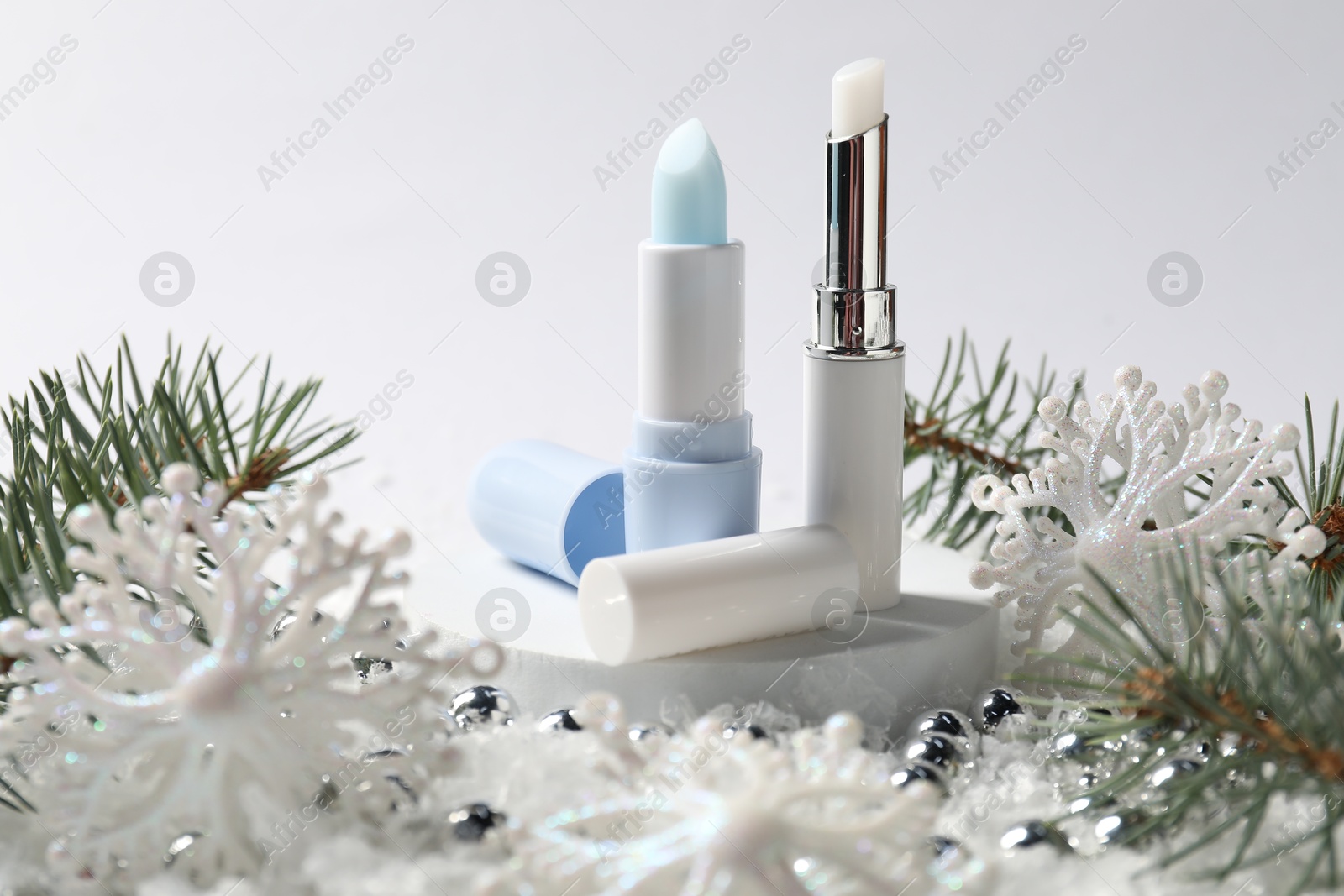 Photo of Moisturizing lip balms, Christmas decor and fir tree branches on artificial snow against white background, closeup. Winter care
