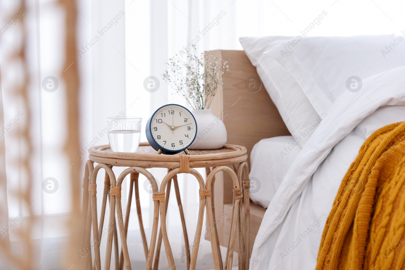 Bedside table with alarm clock, flowers in vase and glass of water near bed indoors Photo of Bedside table with alarm clock, flowers in vase and glass of water near bed indoors