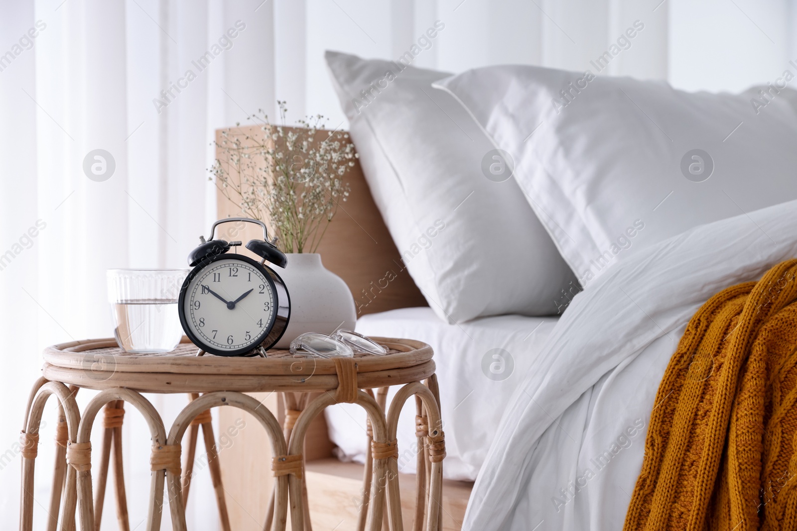 Bedside table with alarm clock, flowers in vase, glass of water and glasses near bed indoors Photo of Bedside table with alarm clock, flowers in vase, glass of water and glasses near bed indoors