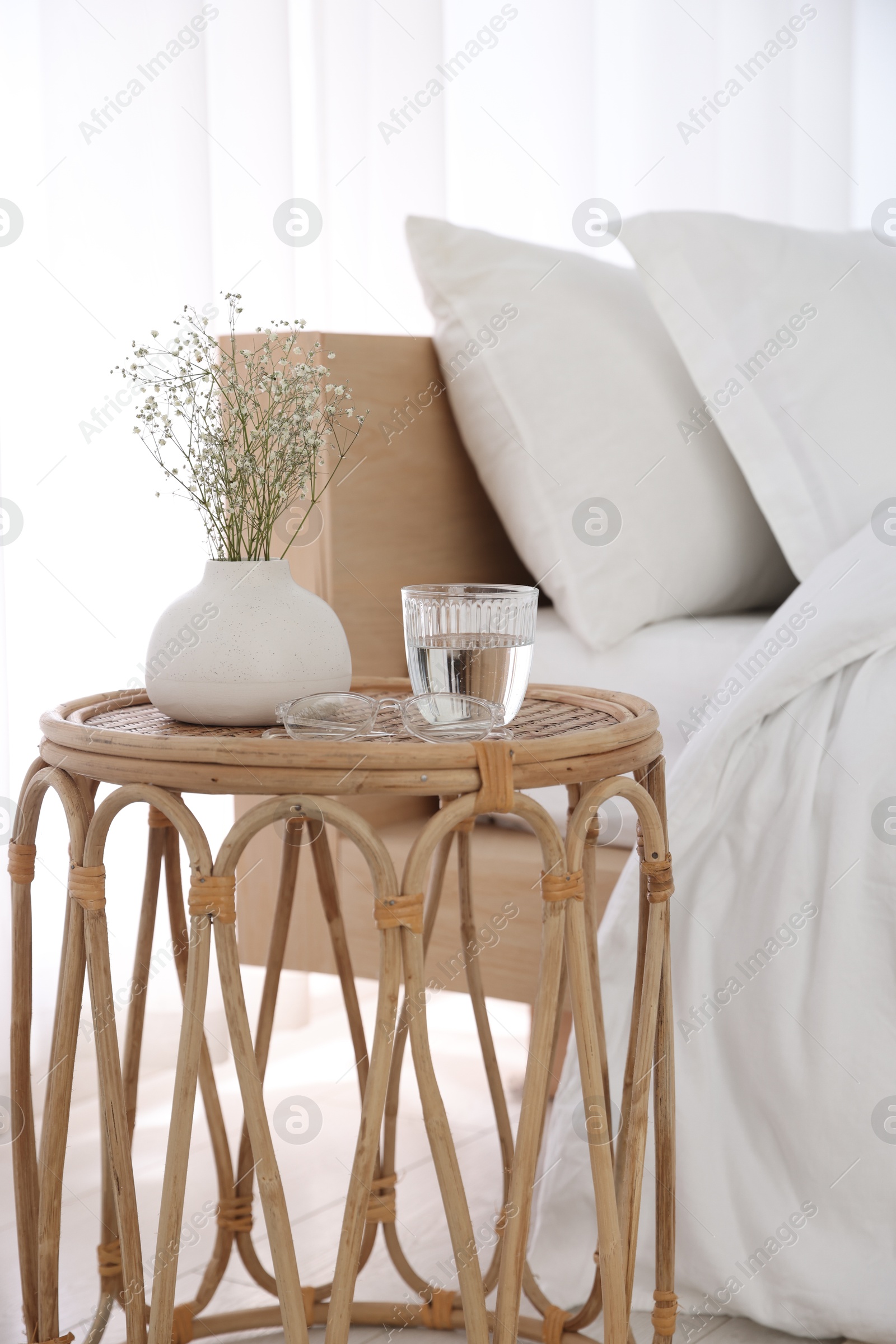 Bedside table with flowers in vase, glass of water and glasses near bed indoors Photo of Bedside table with flowers in vase, glass of water and glasses near bed indoors
