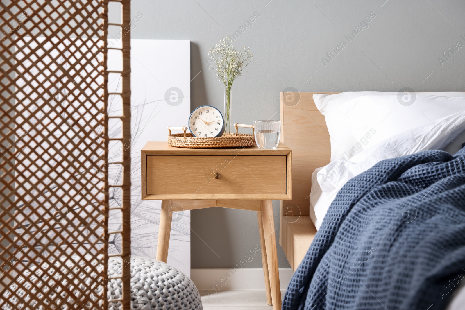 Bedside table with alarm clock, flowers in vase and glass of water near bed indoors Photo of Bedside table with alarm clock, flowers in vase and glass of water near bed indoors
