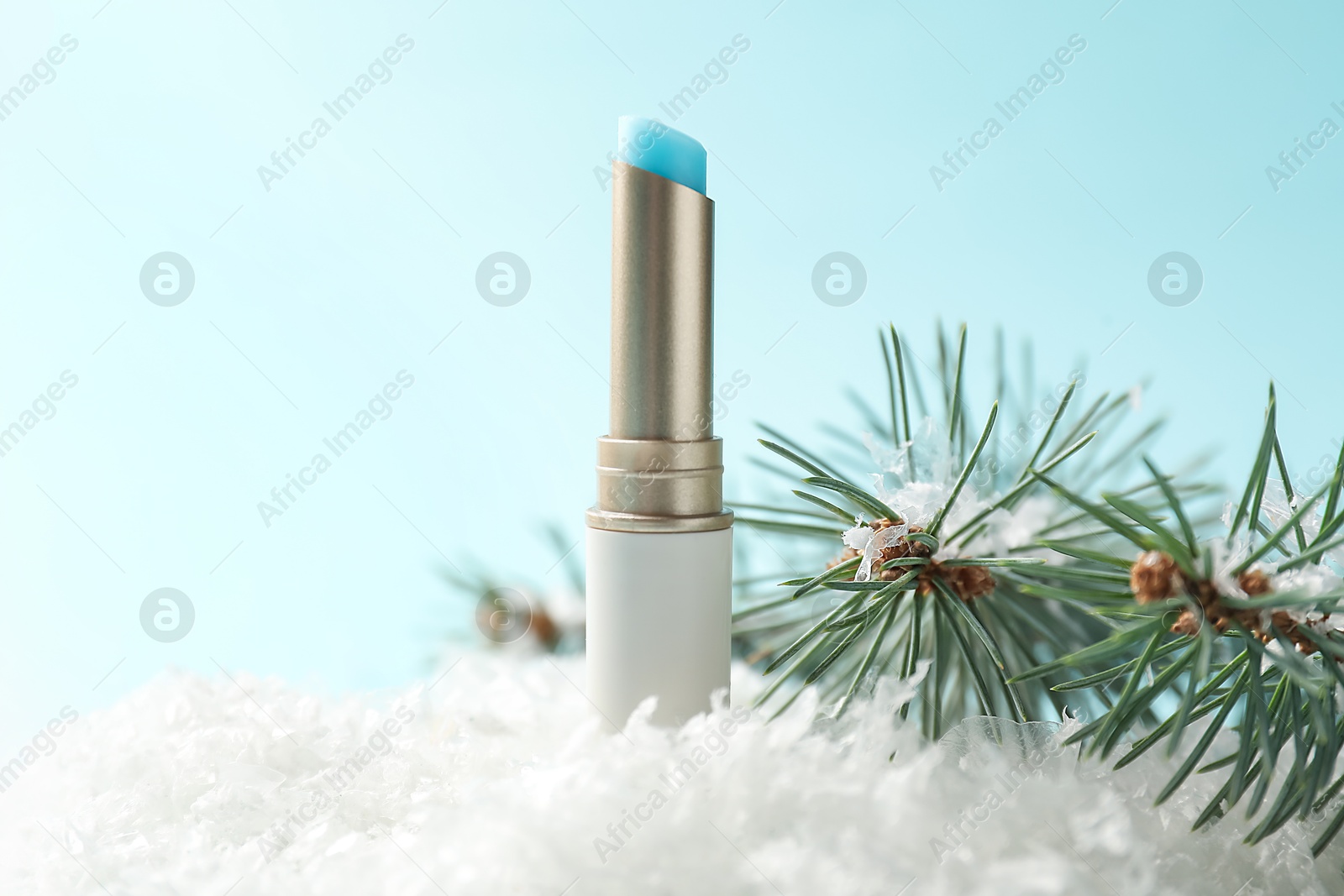 Photo of Moisturizing lip balm and fir tree branches on artificial snow against light blue background, closeup. Winter care