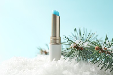 Photo of Moisturizing lip balm and fir tree branches on artificial snow against light blue background, closeup. Winter care