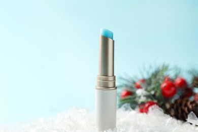 Photo of Moisturizing lip balm and Christmas decor on artificial snow against light blue background, closeup. Winter care