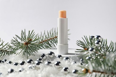 Photo of Moisturizing lip balm, Christmas decor and fir tree branches on artificial snow against white background, closeup. Winter care
