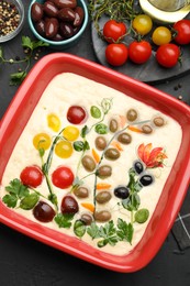 Photo of Raw focaccia with vegetables, olives and spices on black table, flat lay