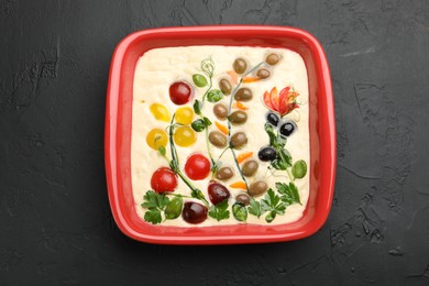 Photo of Raw focaccia with vegetables and olives in baking dish on black table, top view
