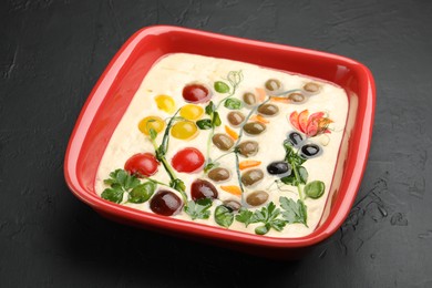 Photo of Raw focaccia with vegetables and olives in baking dish on black table, closeup
