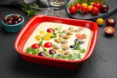 Photo of Raw focaccia with vegetables, olives and spices on black table, closeup