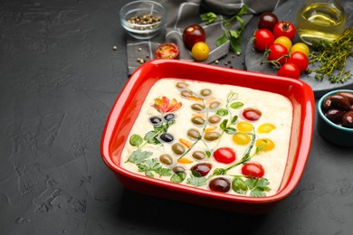 Photo of Raw focaccia with vegetables, olives and spices on black table, closeup