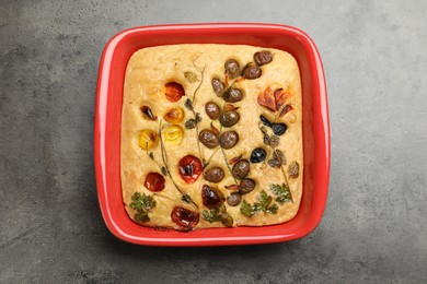 Photo of Delicious focaccia with vegetables and olives in baking dish on grey table, top view