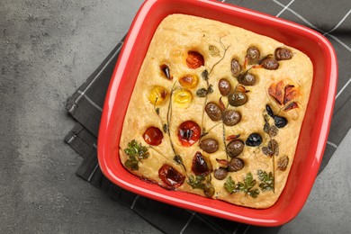 Photo of Delicious focaccia with vegetables and olives in baking dish on grey table, top view. Space for text