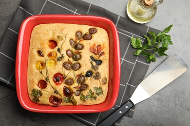 Photo of Delicious focaccia with vegetables, olives, spices and server on grey table, flat lay