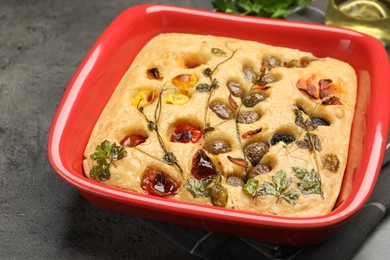 Photo of Delicious focaccia with vegetables and olives in baking dish on grey table, closeup