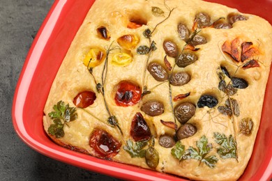Photo of Delicious focaccia with vegetables and olives in baking dish on grey table, above view
