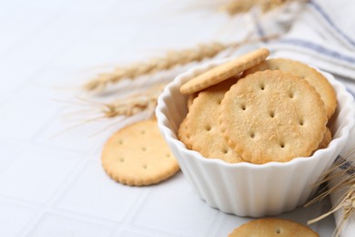 Photo of Tasty salty crackers with wheat spikes on white tiled table, closeup. Space for text