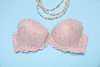 Photo of Beautiful beige lace bra and pearl necklace on light blue background, flat lay