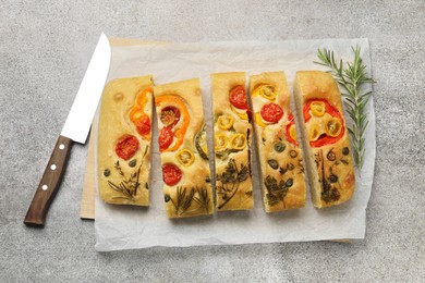 Photo of Cut delicious focaccia with flowers made of vegetables, knife and rosemary on grey table, top view